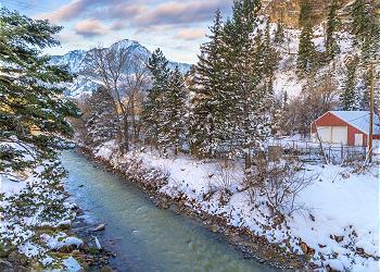 A serene winter scene captures the flowing Uncompahgre River, bordered by snow-covered banks and evergreen trees. Majestic mountains rise in the background, complementing the tranquil atmosphere. A rustic red structure is partially visible along the river, enhancing the natural beauty of the setting.