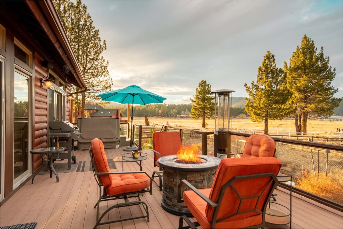A spacious deck features a fire pit surrounded by four lounge chairs in a vibrant orange hue. A gas grill is positioned nearby, along with a shaded umbrella for relaxation. The scenic landscape showcases trees and mountains in the distance, creating a serene outdoor setting.