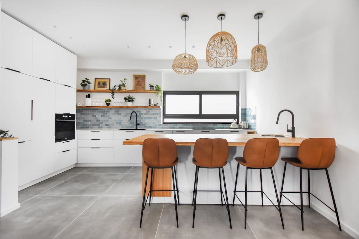 A modern kitchen features sleek white cabinetry and a minimalist design. A wide, wooden countertop is complemented by four contemporary bar stools with brown upholstery. Natural light enters through a large window above the sink, enhancing the fresh and spacious ambiance.