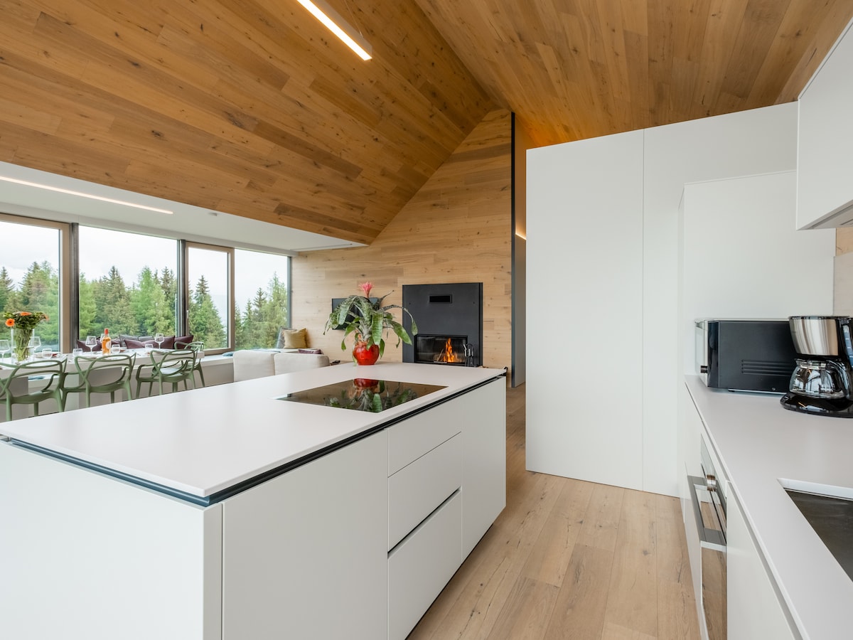 An open kitchen is designed with a large white island that features a cooktop, surrounded by modern cabinetry. Floor-to-ceiling windows allow natural light to illuminate the space, with views of the surrounding greenery. A cozy seating area with a fireplace is visible in the background.