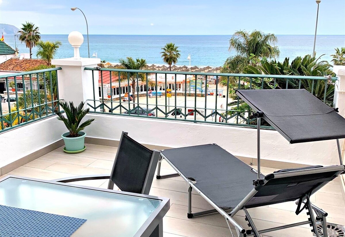 An outdoor terrace features two loungers positioned for relaxation, with a view of the sea. A small table sits nearby, complemented by potted greenery. The scenic backdrop includes palm trees and beach umbrellas along the coastline.