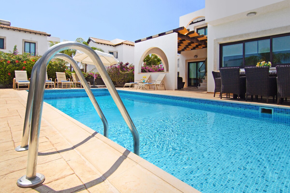A private pool is framed by a spacious patio area, featuring lounge chairs and an outdoor dining set. Colorful vegetation adds a touch of nature, while a shaded area is provided by the covered seating space. Clear skies reflect on the water's surface.
