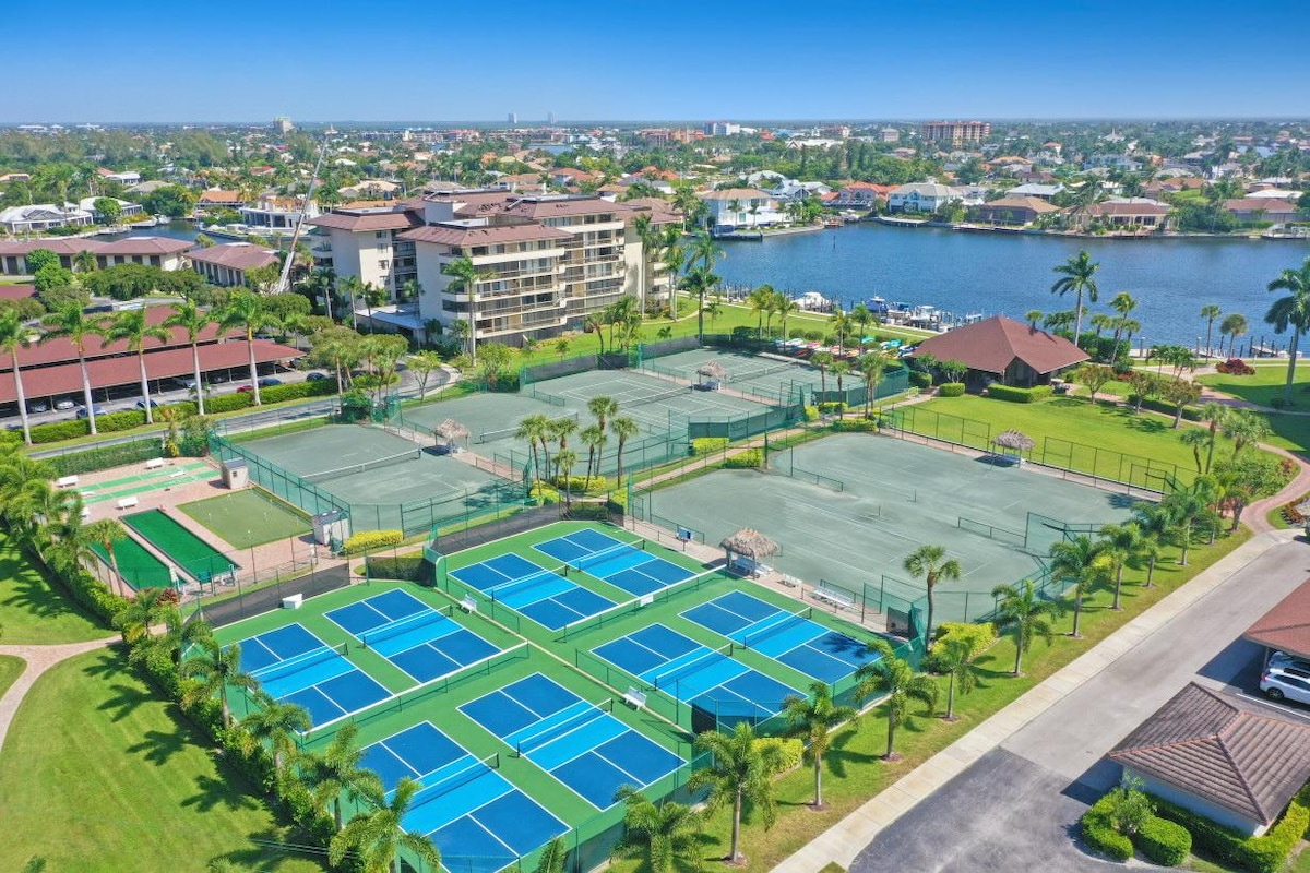 Aerial view of a sports facility featuring multiple tennis and pickleball courts surrounded by palm trees. The layout includes green and blue court surfaces with sunlit areas. Nearby, scenic landscaping and water views create a serene environment, enhancing the recreational experience.