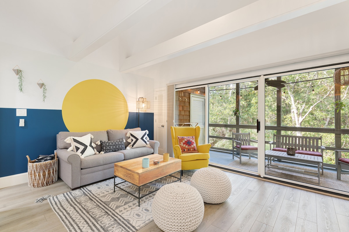 The living area features a comfortable gray sofa paired with a vibrant yellow armchair. A wooden coffee table is centered on a patterned area rug. Expansive sliding glass doors open to a screened porch, providing views of the surrounding greenery.