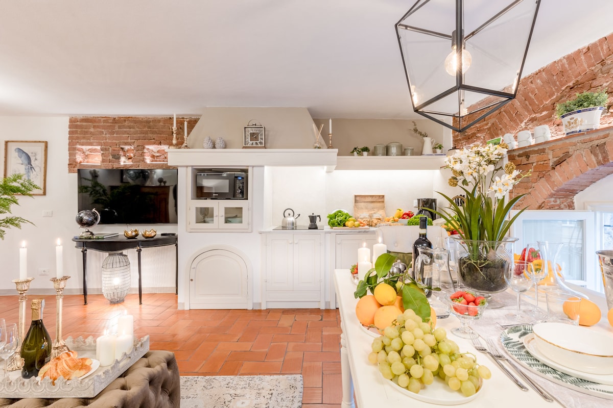 The kitchen area features a mix of modern appliances and rustic charm, highlighted by exposed brick walls. A well-decorated countertop displays fresh fruit, candles, and elegant tableware, while a cozy dining setup is visible, enhancing the inviting atmosphere of the space.