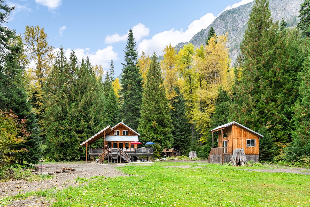 A rustic log cabin and companion bunk house are nestled among tall trees and vibrant fall foliage. A grassy area spreads between the buildings, with outdoor seating present on the deck and a picnic table in the background, all set against a backdrop of mountains.