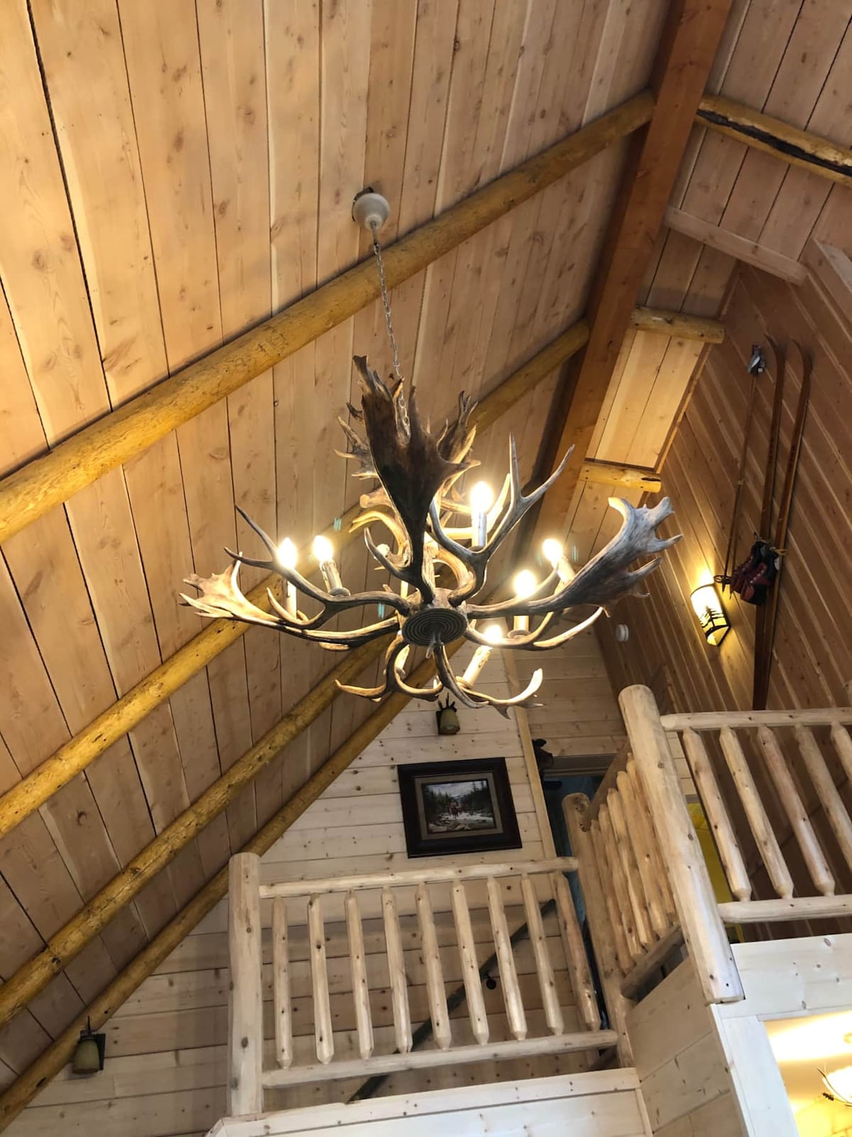 A rustic chandelier made of antlers hangs from a wooden ceiling, providing soft lighting. The vaulted ceiling features exposed beams and natural wood paneling, enhancing the cabin's traditional character. A framed artwork is visible on the wall, adding to the cabin's inviting ambiance.