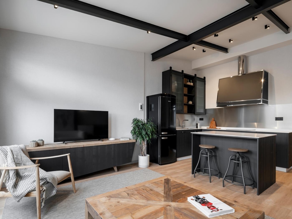 An open-concept kitchen and living area is visible, featuring sleek black cabinetry and modern appliances. A comfortable armchair sits opposite a wooden coffee table, with a plush rug underfoot. Large windows allow natural light to brighten the space, enhancing its welcoming atmosphere.