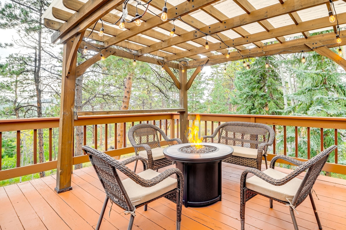 An outdoor deck is surrounded by trees, featuring a wooden pergola with string lights. Four wicker chairs are positioned around a circular fire pit. The warm wooden flooring complements the natural setting, creating a cozy space for relaxation and conversation.