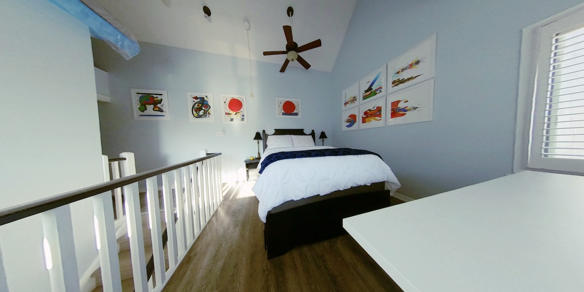 An open loft bedroom features a spacious bed dressed in white linens. Colorful artwork adorns the walls, while a ceiling fan provides ventilation. Natural light filters through the window, illuminating the light blue walls and wooden flooring.
