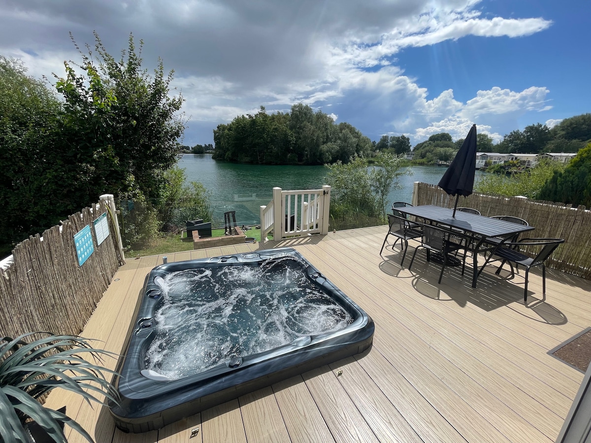 A sunken hot tub is positioned on a spacious deck, overlooking a serene lake surrounded by greenery. A dining table with chairs is available nearby, complemented by an umbrella for shade. The sky features scattered clouds, adding a tranquil ambiance to the view.