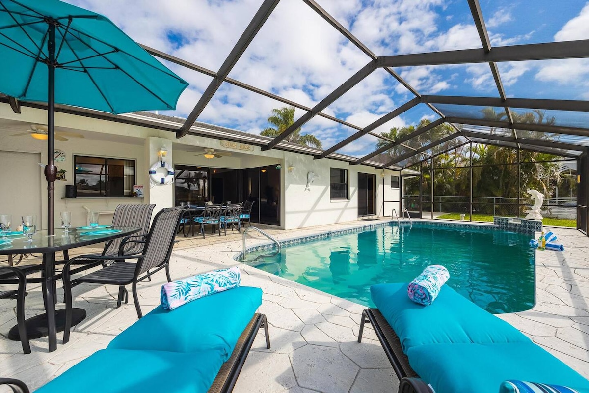 A spacious outdoor area is showcased with a private heated pool surrounded by a screened enclosure. Lounge chairs and a table set are visible, along with a large umbrella offering shade. The tranquil setting features palm trees and a clear sky.