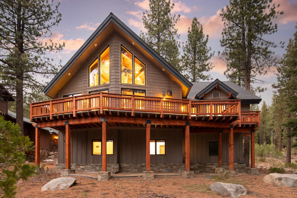A modern chalet features expansive windows that enhance natural light and provide views of the surrounding trees. The wooden deck is adorned with rustic railings, inviting outdoor relaxation. The home's structure showcases a combination of wood and stone, set against a scenic backdrop of dusk-lit foliage.