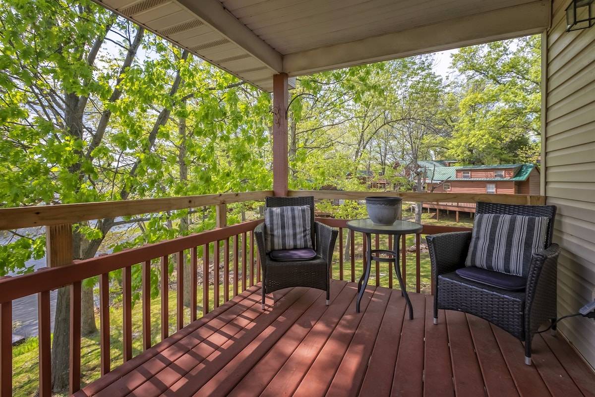 A shaded outdoor deck features two wicker chairs with cushions and a small round table, surrounded by lush greenery. The wooden flooring contrasts with the natural elements, creating a cozy space for relaxation. Nearby, glimpses of the lake can be seen through the trees.