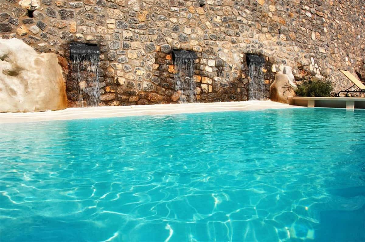 A clear blue pool is framed by a stone wall featuring three small waterfalls cascading gently. The water reflects the sunlight, creating a serene atmosphere in this shared outdoor space.