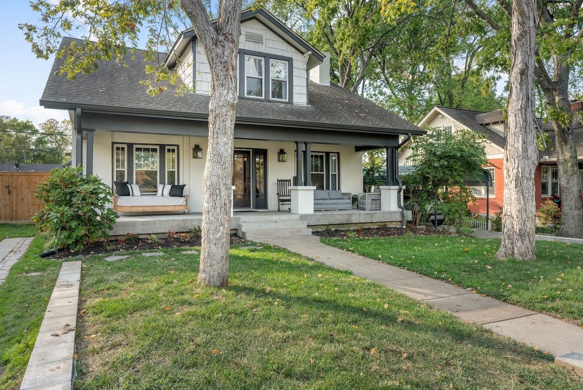 The exterior of a charming two-story Craftsman home is displayed, featuring a welcoming front porch with seating. Lush green grass and trees surround the property, while a concrete walkway leads to the entrance. The home presents a blend of traditional architecture and modern design.