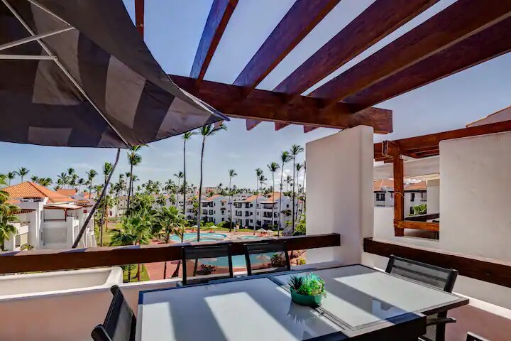 A spacious balcony is depicted, featuring a dining table with chairs shaded by an umbrella. Views of the surrounding palm trees and pool area can be seen, with residential buildings in the background, showcasing the outdoor space's inviting atmosphere.