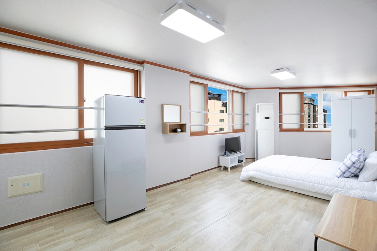 A spacious room is featured, with natural light filtering through large windows. A soft mattress is placed on the floor, accompanied by decorative pillows. A refrigerator, television, and a simple table create a functional living space, while light walls enhance the overall brightness.