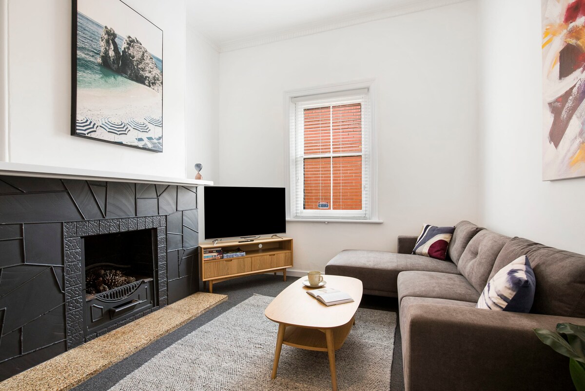 A spacious living area is presented with a large corner lounge suite and a flat screen TV placed on a wooden console. A decorative fireplace enhances the space, while natural light enters through a window with red accents. A coffee table occupies the center, creating a cozy atmosphere.