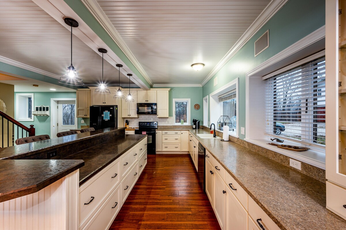 The spacious kitchen features light-colored cabinetry and a large countertop that extends to both sides. Multiple pendant lights provide illumination. Modern appliances, including a stove and microwave, are visible. Windows allow natural light to brighten the room, enhancing the functional layout.