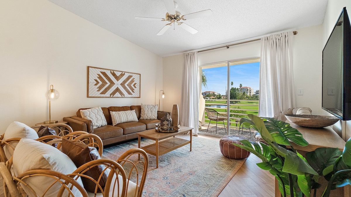 A spacious living area is displayed, featuring a comfortable brown sofa and two rattan chairs arranged around a wooden coffee table. Large glass doors open to a picturesque view of the golf course, complemented by light, airy curtains and a potted plant in the corner.