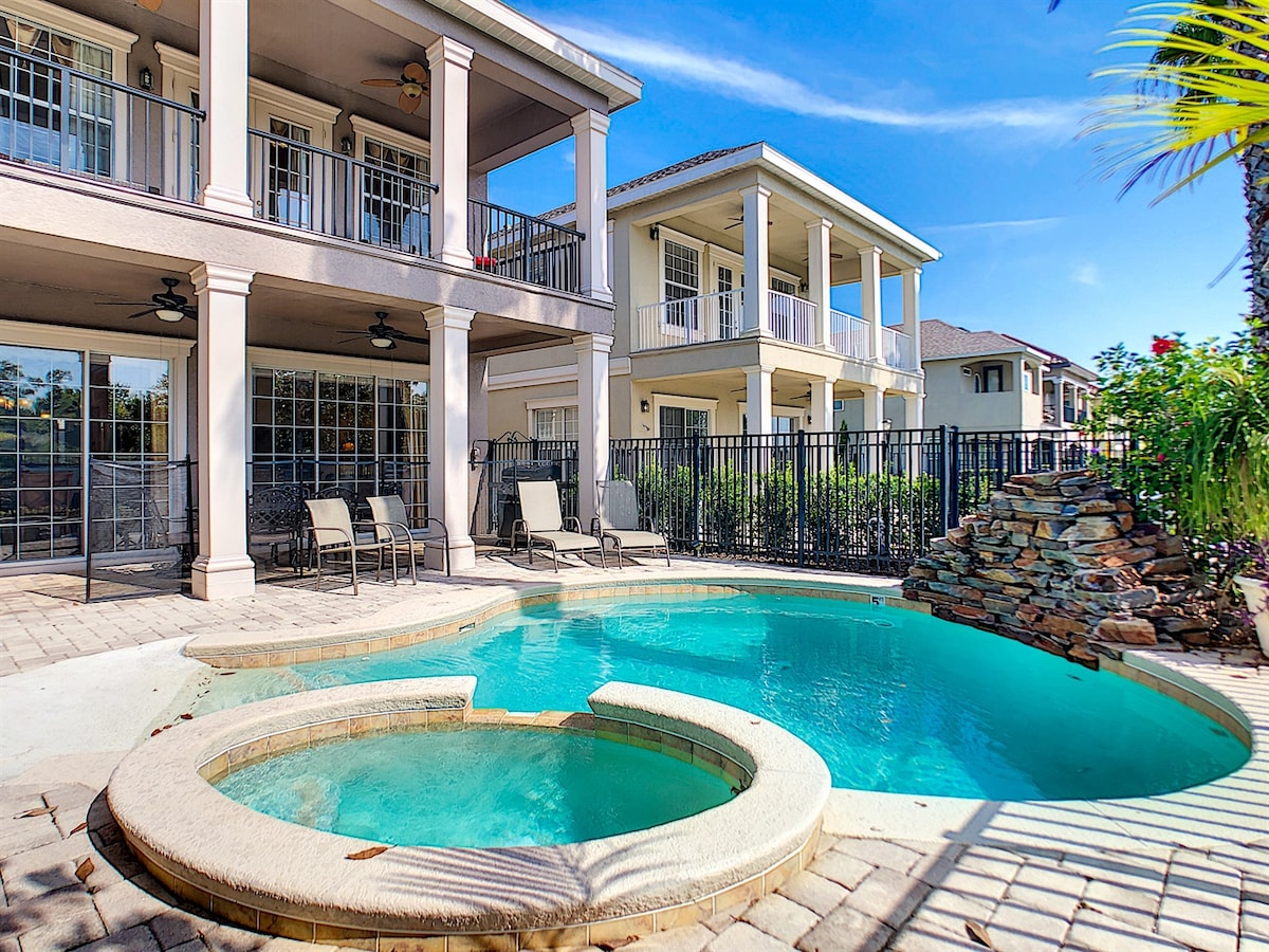 A private pool area is featured, showcasing a curved spa attached to a turquoise pool. Surrounding the pool, four lounge chairs are arranged on a stone patio, with a fenced backyard visible in the background. The two-story villa is partially seen, highlighting its expansive balconies.