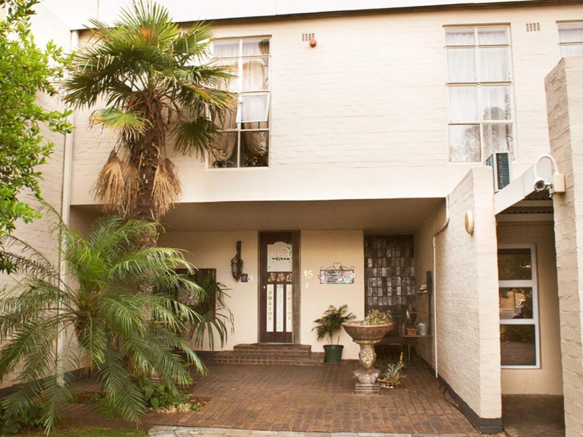 A two-story building exterior features a welcoming entrance with steps leading up to the front door. Lush green palm trees and planters are visible, creating a serene outdoor environment. Large windows provide ample natural light, enhancing the inviting appearance.