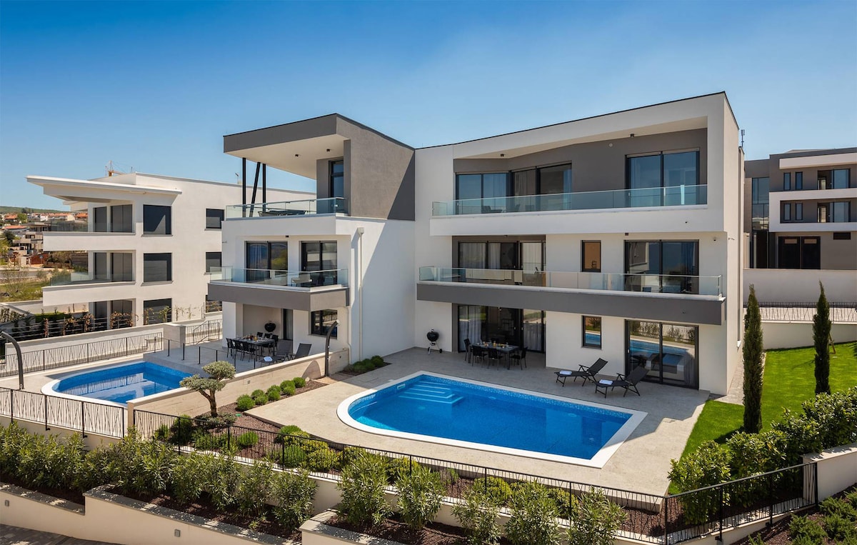 A modern apartment complex is showcased, featuring multiple levels and large balconies. Two private pools are positioned at the forefront, surrounded by landscaped greenery. Sun loungers are arranged near the pools, creating a relaxing environment for guests.