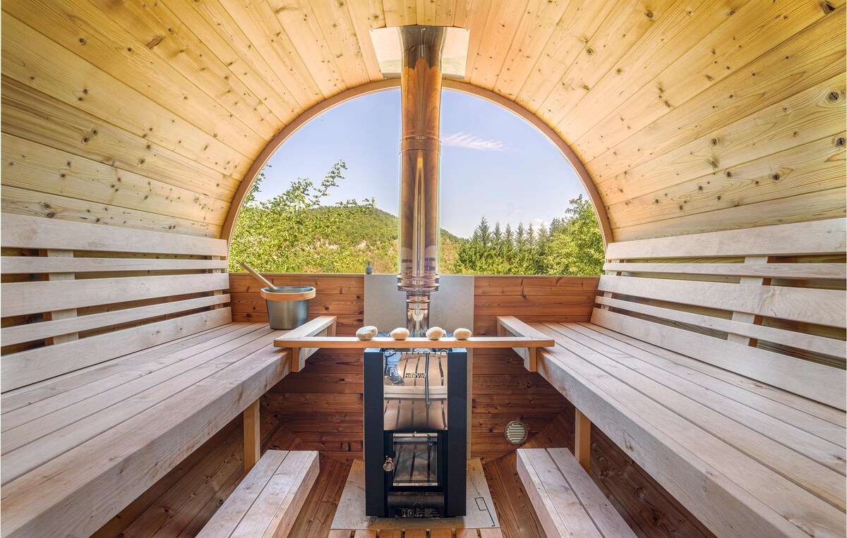 The interior of the sauna features smooth wooden benches arranged along the curved walls. A central stove is visible, surrounded by a minimalist table setting. Natural light filters in through the arching structure, providing a warm ambiance and views of the surrounding greenery.