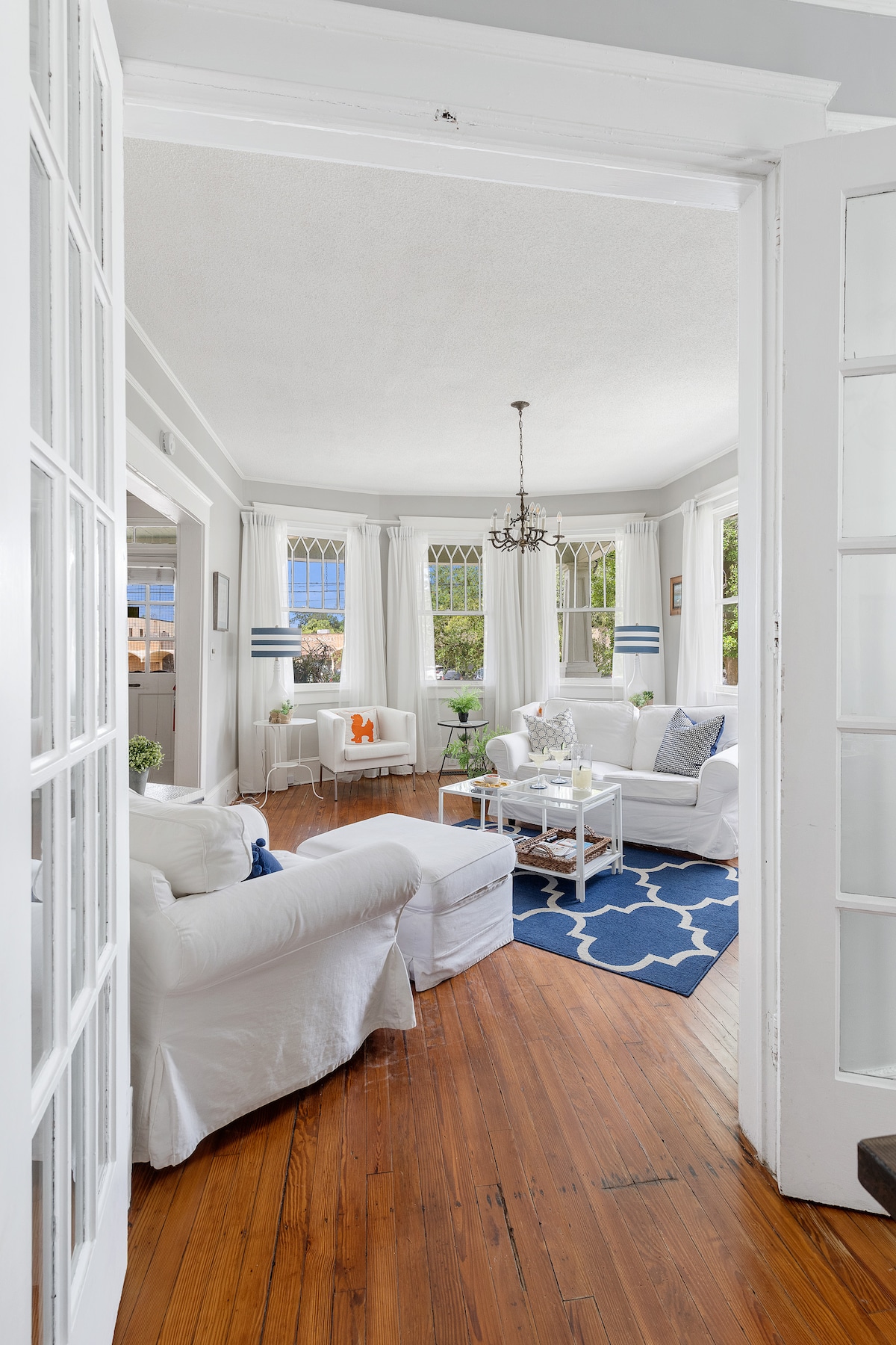 The living area features a bright and airy atmosphere created by large windows draped with sheer curtains. Plush white sofas are arranged around a coffee table, complementing the stylish blue area rug. A chandelier hangs from the ceiling, enhancing the inviting space.