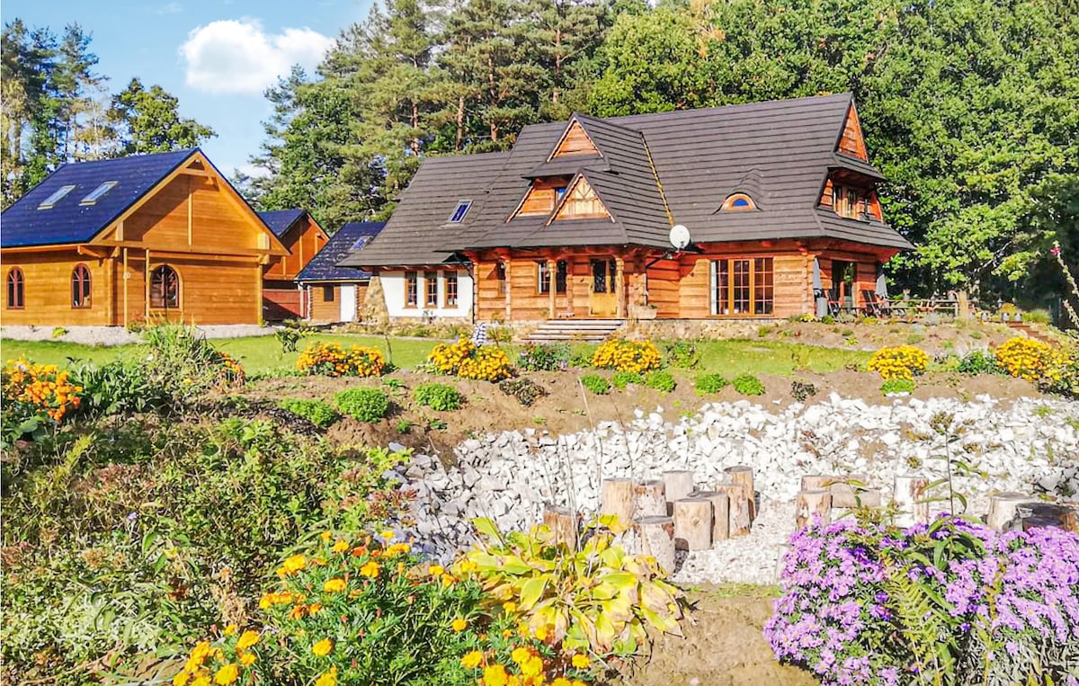 A charming wooden house is framed by colorful flower beds. The property features a spacious patio, large windows, and a traditional roof design. Surrounding trees offer a natural backdrop, while nearby structures enhance the cozy setting of this inviting retreat.