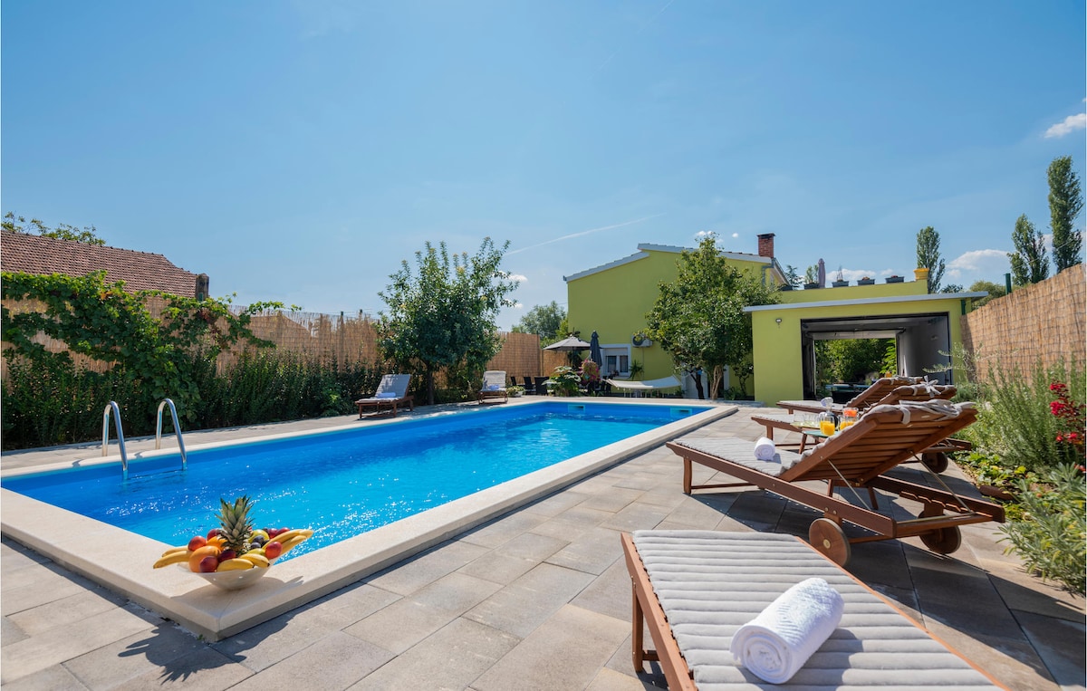 A private swimming pool offers a refreshing escape, surrounded by greenery and smoothly paved patio areas. Lounge chairs are arranged beside the pool, accompanied by fruit displays. Architectural features of the house and outdoor dining space are visible in the background under clear skies.