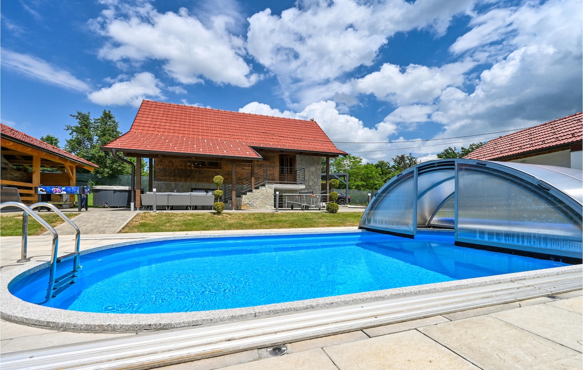 A clear swimming pool is situated in the foreground, surrounded by stone tiles. The house with red-tiled roofs and wooden accents can be seen in the background, accompanied by a covered terrace area and green landscaping. The sky is partly cloudy, enhancing the outdoor setting.