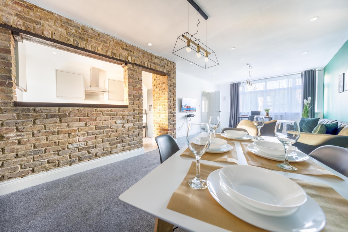An open-plan dining area is highlighted by a stylish table set for six, featuring plates and glasses. A textured brick wall adds warmth to the space, while a modern light fixture hangs above. The inviting living area is visible in the background, complemented by large windows.