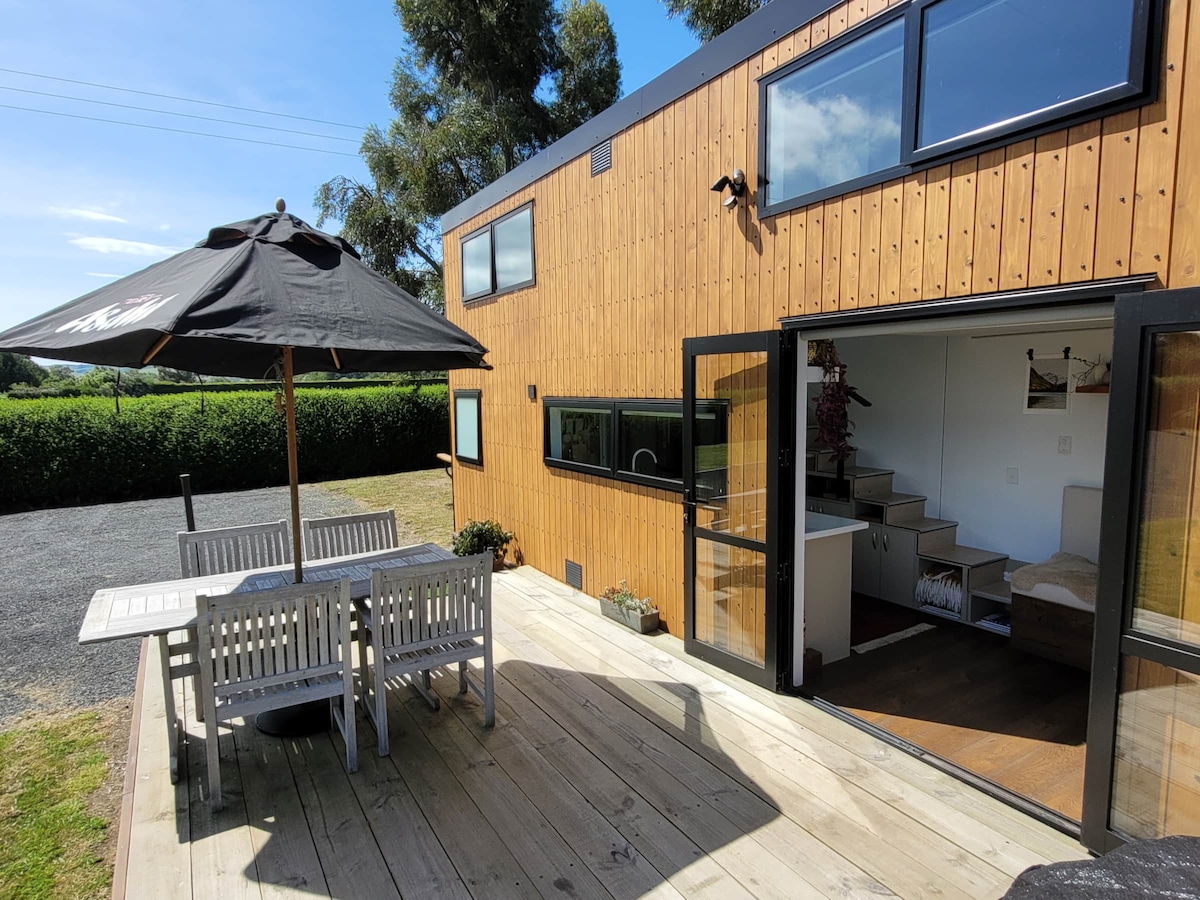 An outdoor deck area features a wooden dining table with matching chairs and a large umbrella providing shade. The two-story tiny home is constructed with wood paneling, and large windows illuminate the space. Lush greenery surrounds the property, contributing to a calm setting.