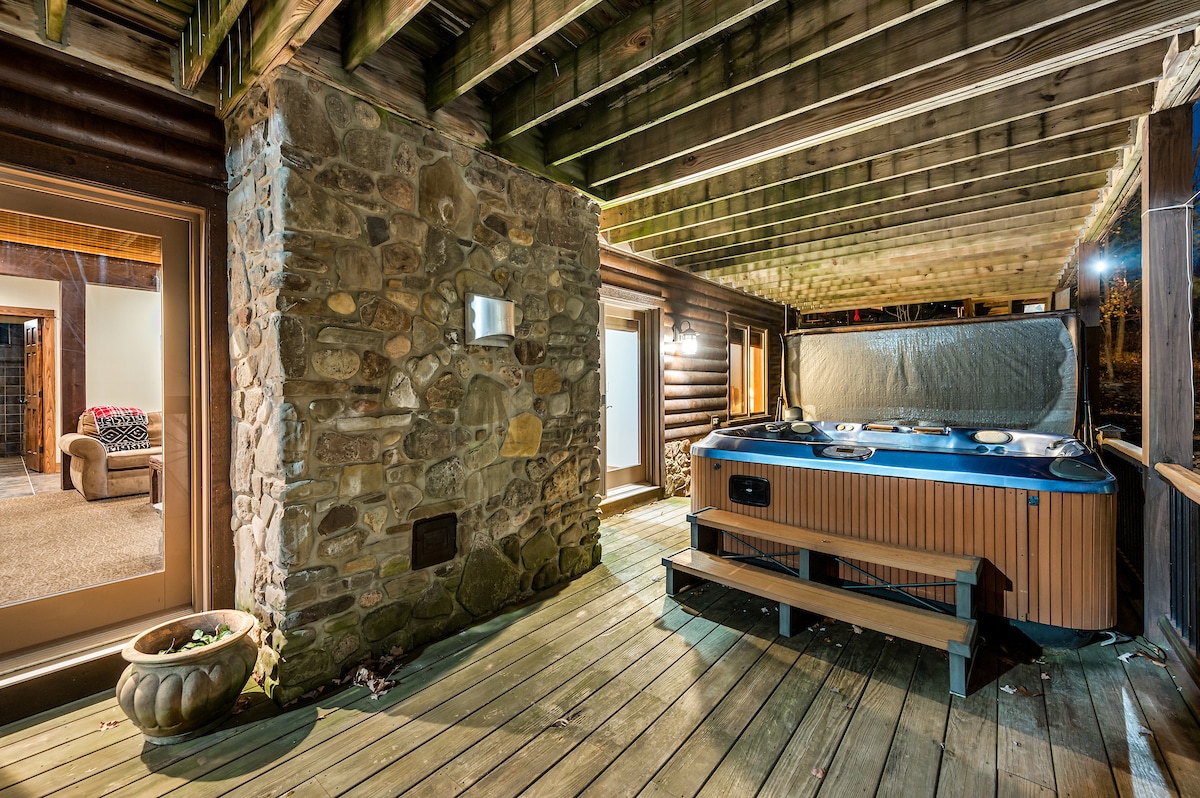 An outdoor area features a covered hot tub surrounded by wooden decking. Stone walls add rustic charm, while a cozy seating area is seen in the background. Warm lighting highlights the space, creating an inviting atmosphere for relaxation.
