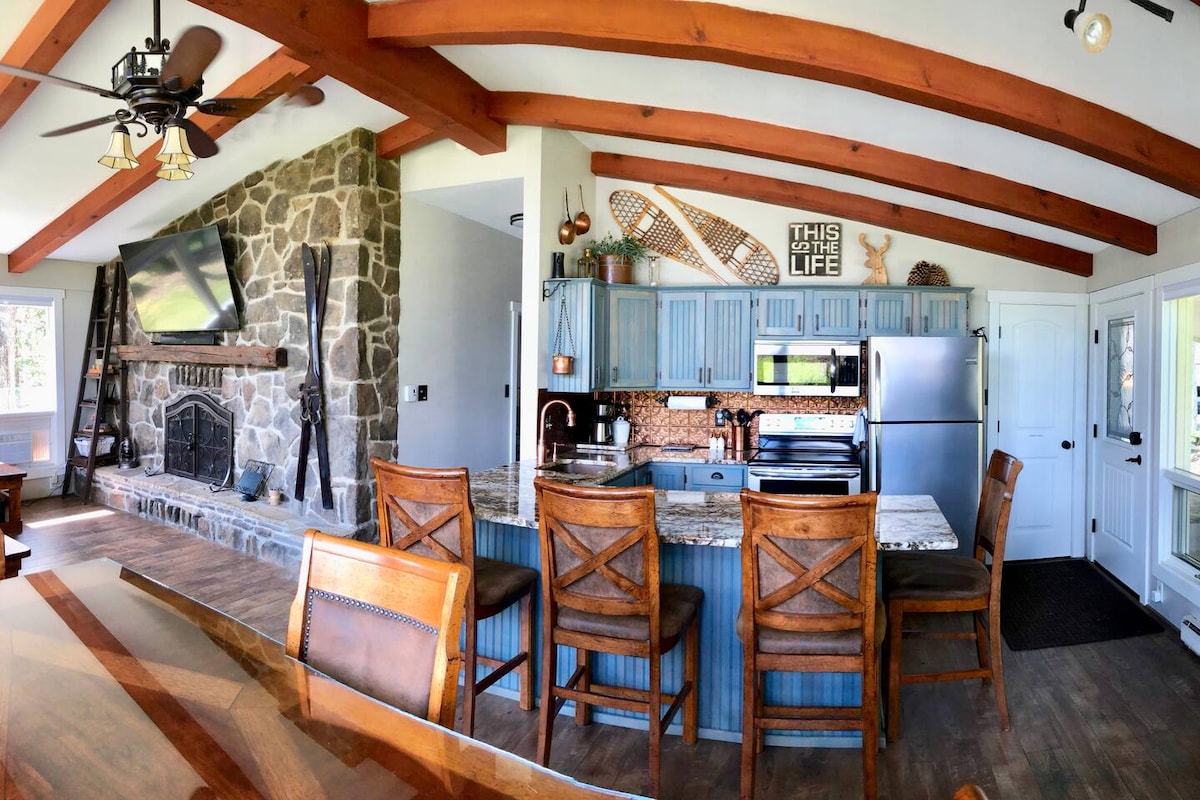 The kitchen features custom granite countertops and stainless steel appliances, surrounded by blue cabinetry. A spacious island with four wooden stools provides seating. The stone fireplace adds character to the open layout, complemented by exposed wooden beams overhead and ample natural light from the windows.
