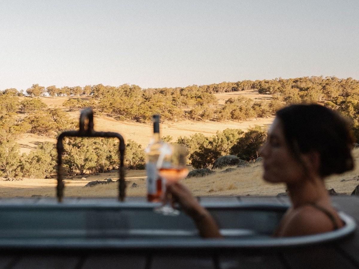 A person enjoys a glass of rosé while relaxing in a bathtub, with vast, open landscapes in the background. Rolling hills and scattered trees create a serene natural setting, enhancing the tranquil atmosphere of the outdoor bath.