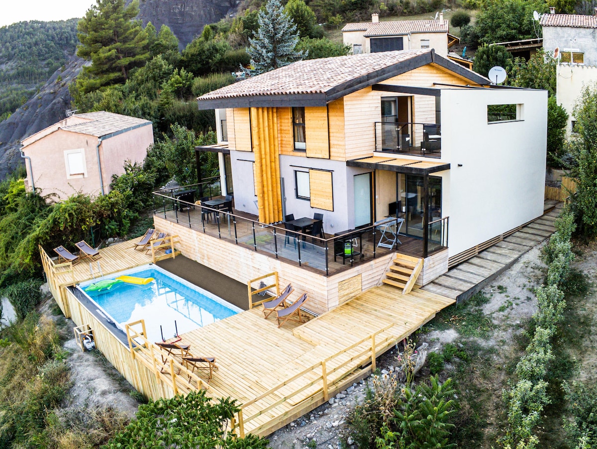 A modern two-story building is situated on a hillside, showcasing a wooden deck surrounding a rectangular pool. Glass railings provide a clear view of the outdoor area, which includes seating and sun loungers. Natural vegetation frames the property, enhancing its ecological design.