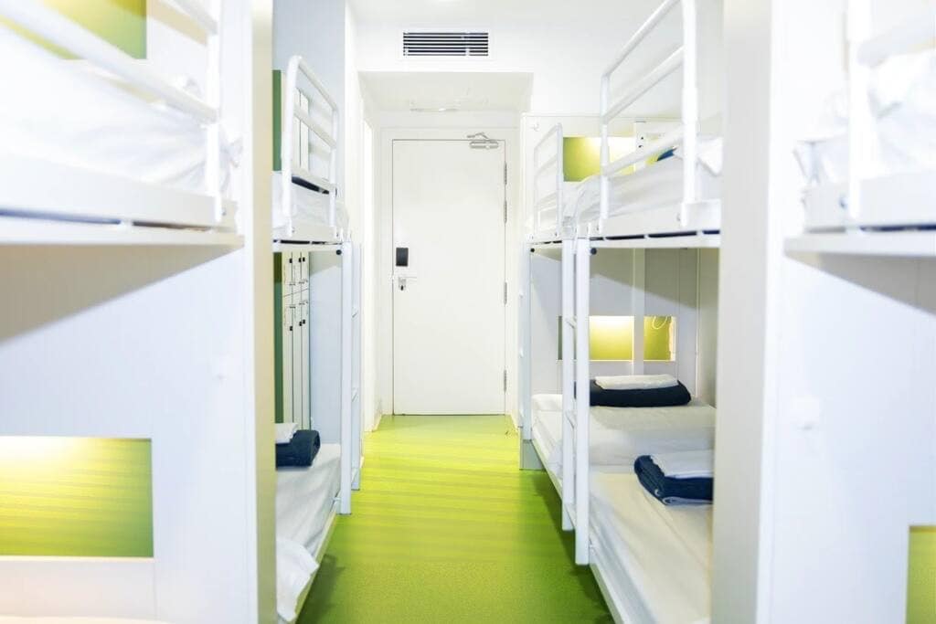 A bright and airy dormitory space features multiple bunk beds arranged along the walls. Each bed includes a soft mattress, a small reading light, and neatly folded blankets. The floor is covered in a vibrant green carpet, and a door is visible at the far end of the room.
