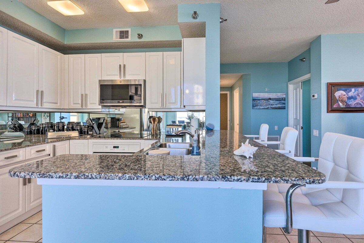 A modern kitchen is showcased, featuring white cabinetry and sleek stainless steel appliances. The granite countertop extends into a breakfast bar with contemporary stools. Mirrors reflect natural light, enhancing the airy feel of the space. The kitchen is fully equipped for cooking and dining needs.