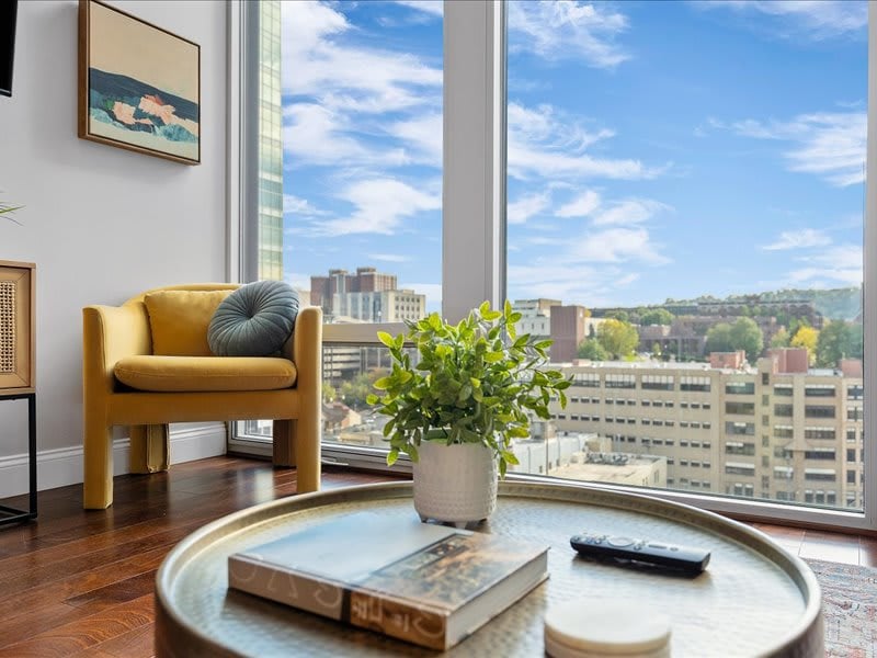 A bright living area is highlighted by a yellow chair positioned near a large window, offering expansive views of the cityscape. A small, round table displays a book and remote, while a potted plant adds a touch of greenery to the space.