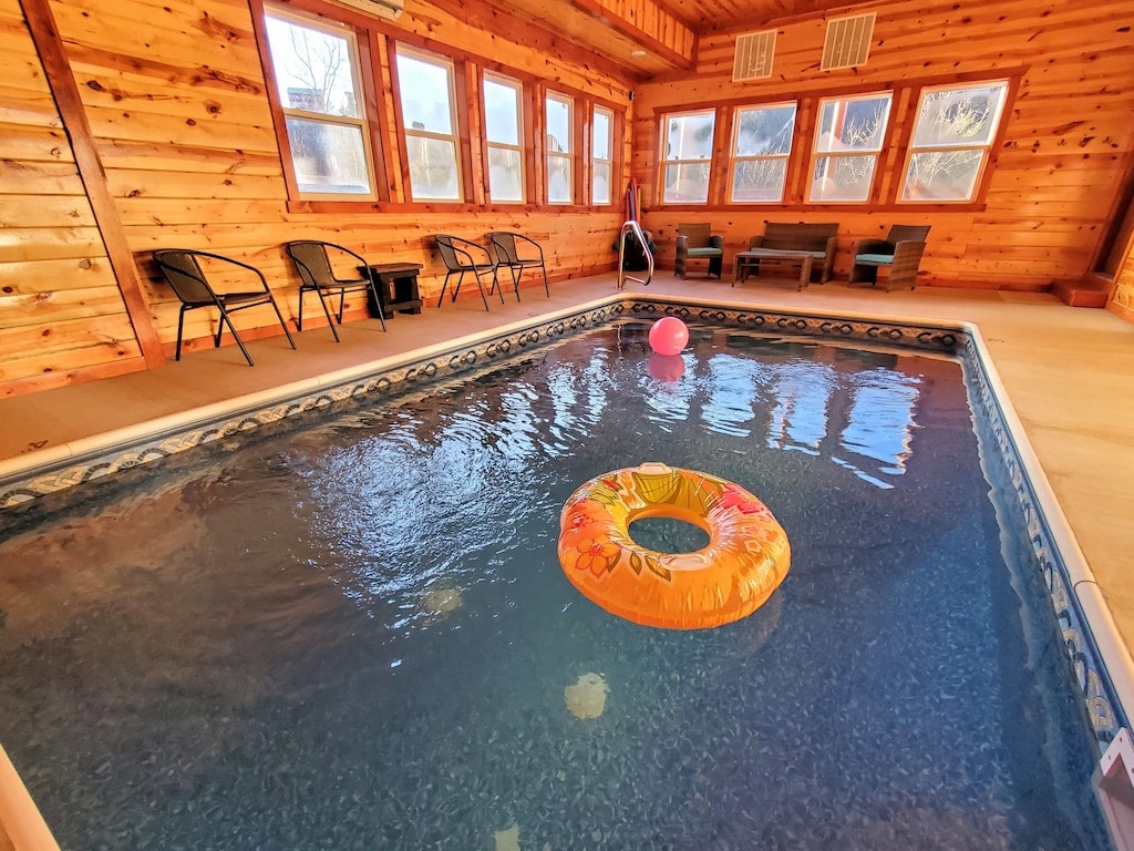 An indoor pool area is surrounded by wooden walls and large windows allowing natural light to fill the space. A colorful inflatable float and a pink ball rest on the water's surface, while poolside seating includes black chairs and cushioned loungers.