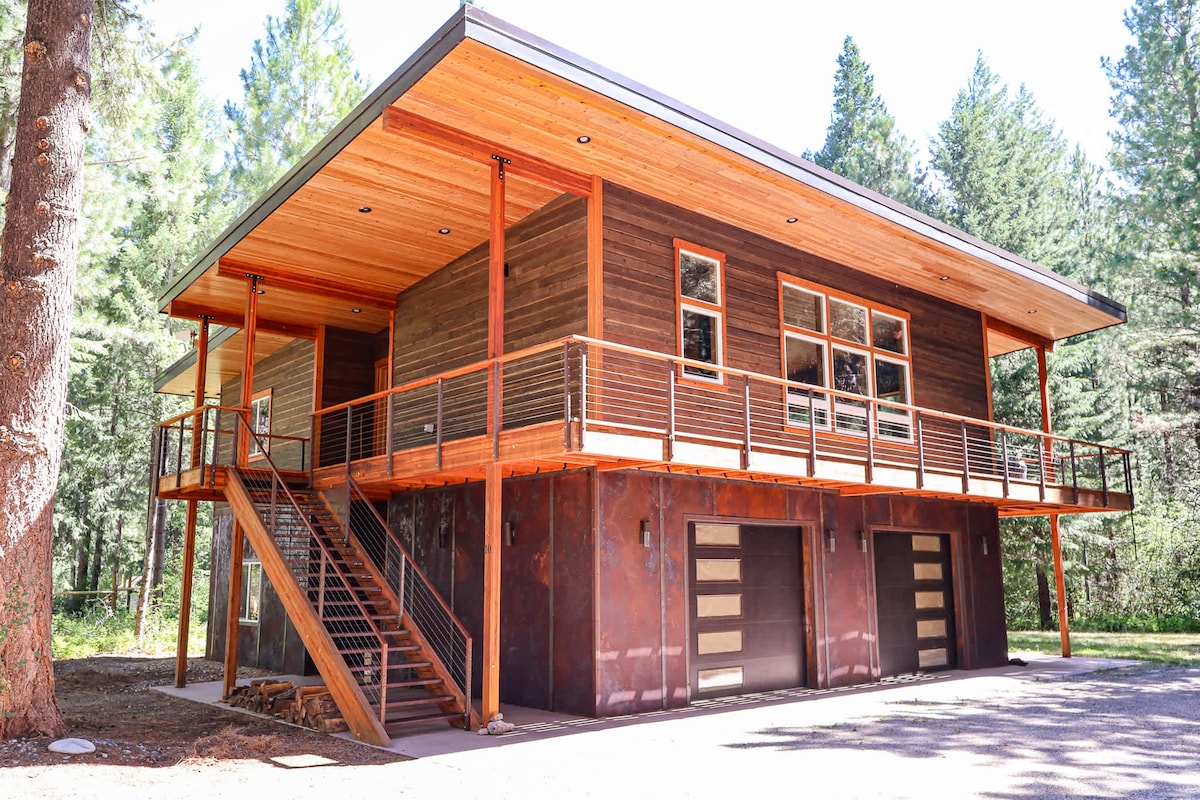 The modern cabin features a wooden exterior with large windows allowing ample natural light. A covered porch extends along the front, with a staircase leading to the elevated entrance. The lower level includes two garage doors, seamlessly integrated into the design.