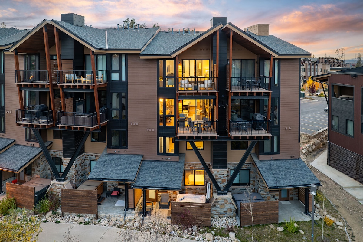 A multi-level townhome exterior is showcased, featuring a blend of wood and stone materials. Balconies are adorned with outdoor furniture, and interior lights are visible through expansive windows. The backdrop features a colorful sunset illuminating the sky, creating a welcoming atmosphere.