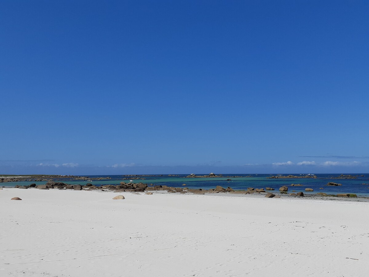 A serene beach scene is displayed, showcasing a wide expanse of soft, white sand meeting clear blue waters. Rocky formations can be seen just offshore beneath a cloudless sky, highlighting the beauty of the coastal environment.