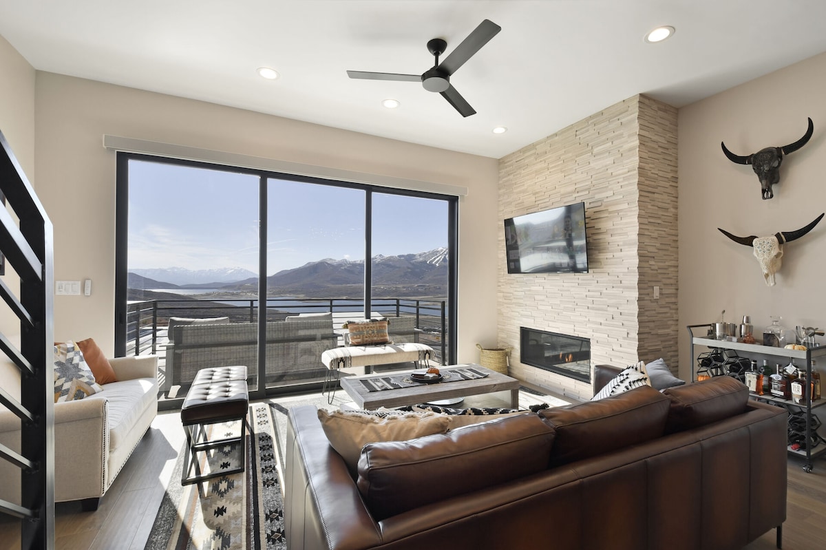 A spacious living area is oriented towards large glass doors that open to panoramic mountain views. A leather sofa sits comfortably, accompanied by a stylish coffee table and an area rug. A stone fireplace adds warmth, while a ceiling fan circulates air.