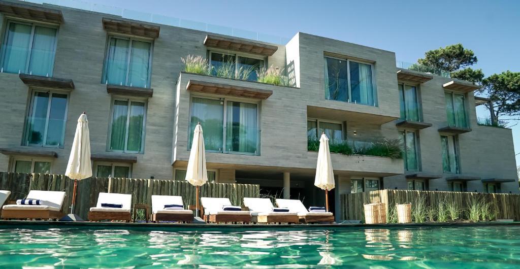 The exterior view showcases a modern building with large windows, surrounded by a serene pool area. Sun loungers with umbrellas line the pool's edge, and lush greenery accents the surrounding landscape, adding to the tranquil environment.