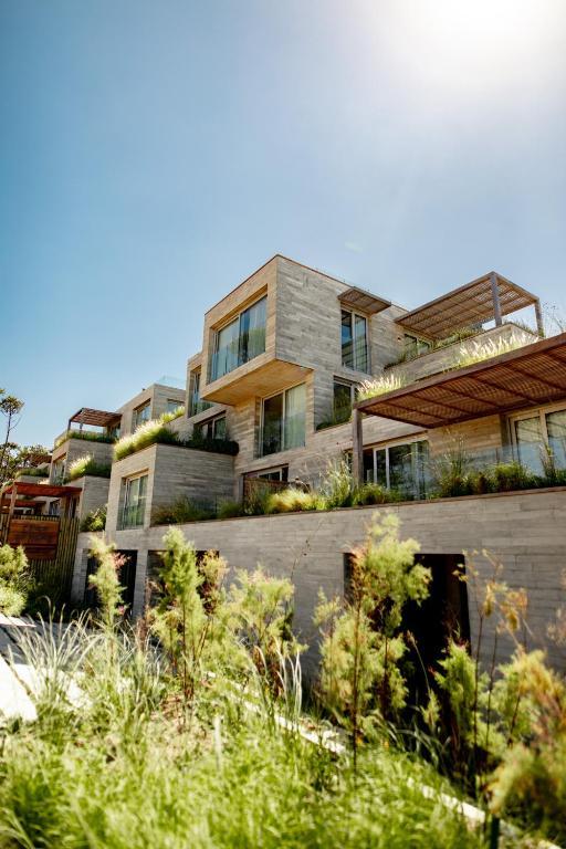 A modern multi-level building is depicted, featuring a blend of stone and wooden elements. Large glass windows are visible, allowing for ample natural light. Native plants and landscaping surround the structure, creating a connection with the natural environment.