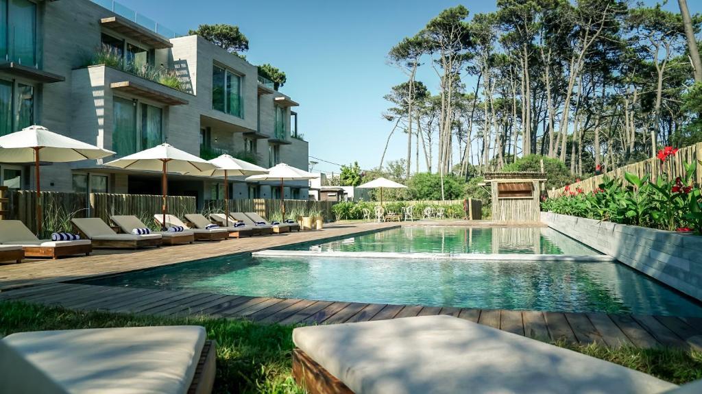 A serene pool area is surrounded by comfortable sun loungers and umbrellas. Lush greenery and tall pine trees provide a natural backdrop, while the calm water reflects the clear sky. The pool features a wooden deck, enhancing the outdoor leisure space.
